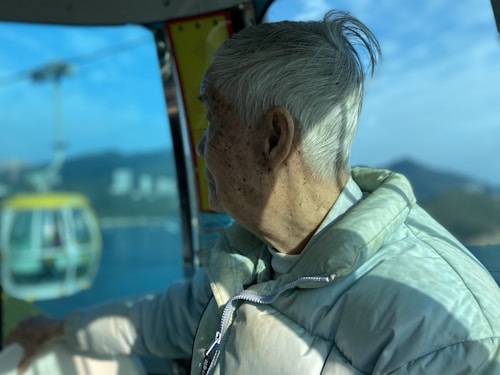 An elderly person with white hair is sitting inside a cable car, wearing a light-colored puffer jacket. The view outside shows a cable car moving along its line, with a scenic backdrop of mountains and a blue sky.