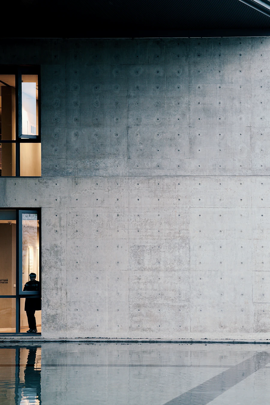 A single figure standing in the deep window reveal of a board-marked concrete facade