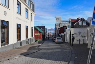 A quaint urban street flanked by various buildings, including a large cream-colored structure with multiple windows. There are colorful houses, one painted red with green accents, and a street lined with parked cars. In the background, distant snow-capped mountains are visible under a clear blue sky.