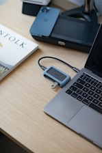 A minimalist workspace features a sleek laptop on a light wooden desk. Nearby, there’s a smartphone and a book with a visible title next to a simple electronic device connected with a cable. The setting is clean, organized, and modern.