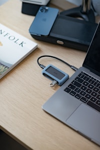 A minimalist workspace features a sleek laptop on a light wooden desk. Nearby, there’s a smartphone and a book with a visible title next to a simple electronic device connected with a cable. The setting is clean, organized, and modern.