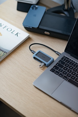 A minimalist workspace features a sleek laptop on a light wooden desk. Nearby, there’s a smartphone and a book with a visible title next to a simple electronic device connected with a cable. The setting is clean, organized, and modern.