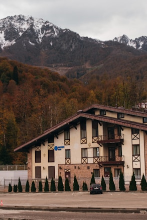 A large chalet-style building with a pitched roof is nestled at the base of a mountainous landscape. The snow-capped peaks stand tall in the background, while the forest surrounding the building displays autumnal foliage. A row of trees is planted along a fence in front of the building, and a lone car is parked in an empty lot.
