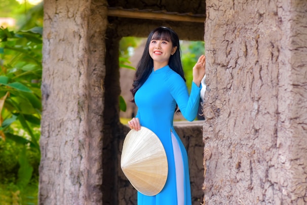 A woman in a bright blue traditional dress stands next to a textured, rustic wall. She is holding a conical hat and looking upward with a smile. The background features lush green foliage, adding a touch of nature to the scene.