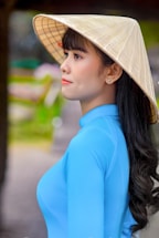 A woman in a light blue traditional Vietnamese ao dai, wearing a conical hat. The background is slightly blurred, with hints of greenery and what appears to be a park setting.