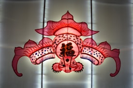 A vibrant, intricately designed kite with symmetrical patterns and bold colors, featuring traditional Asian art motifs and Chinese characters at the center. The kite is predominantly red with hints of pink and white, and has elaborate line work and patterns.