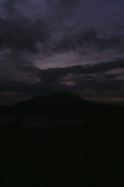 Dark, moody landscape featuring a silhouetted mountain against a cloudy sky. The horizon shows faint hints of light, suggesting dawn or dusk. The scene conveys an atmospheric and serene quality.