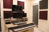 A home studio setup with two electronic keyboards on a wooden desk, featuring a large monitor and two speakers on either side. Acoustic foam panels in maroon and black are mounted on the cream-colored walls. Various electronic devices and a framed photograph are placed on the desk.