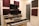 A home studio setup with two electronic keyboards on a wooden desk, featuring a large monitor and two speakers on either side. Acoustic foam panels in maroon and black are mounted on the cream-colored walls. Various electronic devices and a framed photograph are placed on the desk.