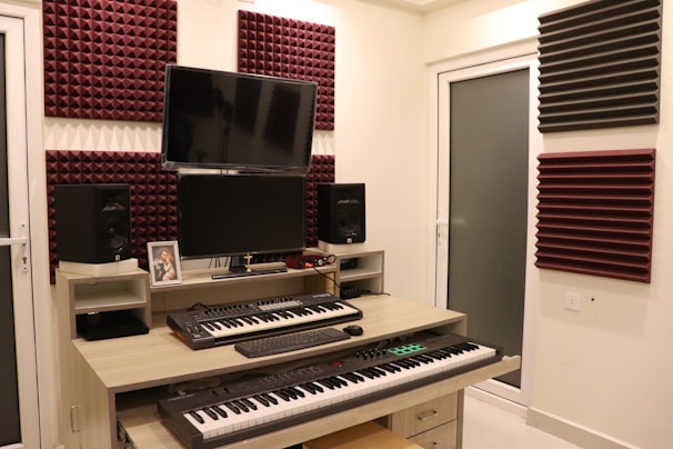A home studio setup with two electronic keyboards on a wooden desk, featuring a large monitor and two speakers on either side. Acoustic foam panels in maroon and black are mounted on the cream-colored walls. Various electronic devices and a framed photograph are placed on the desk.