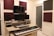 A home studio setup with two electronic keyboards on a wooden desk, featuring a large monitor and two speakers on either side. Acoustic foam panels in maroon and black are mounted on the cream-colored walls. Various electronic devices and a framed photograph are placed on the desk.