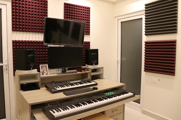A home studio setup with two electronic keyboards on a wooden desk, featuring a large monitor and two speakers on either side. Acoustic foam panels in maroon and black are mounted on the cream-colored walls. Various electronic devices and a framed photograph are placed on the desk.