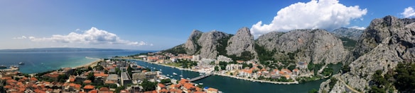 A coastal town nestled between rugged mountains and the sea, with a river winding through. The landscape features a blend of natural beauty with dense rocky cliffs and lush greenery. The architecture includes a mix of terracotta-roofed buildings and modern structures.