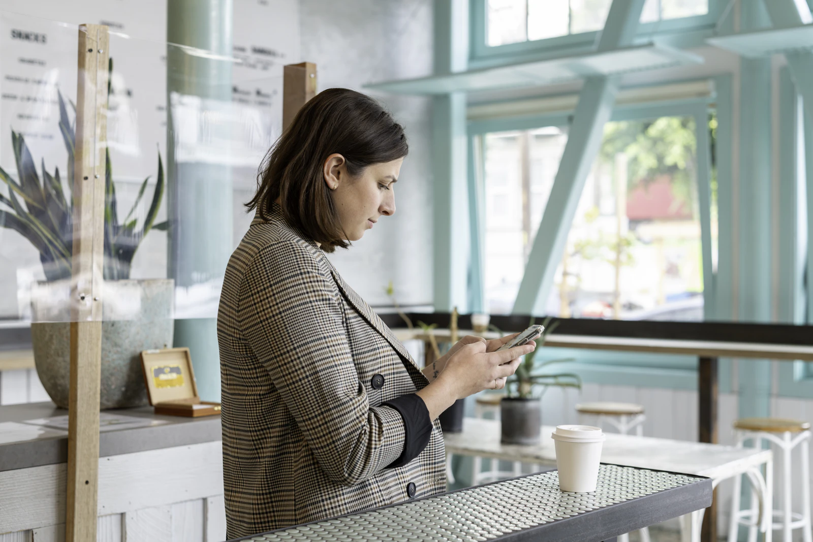 A woman in a checked shirt checking her smartphone while standing at a cafe counter