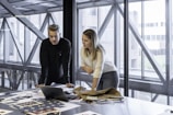 Two people are focused on a laptop, surrounded by papers and fabric samples on a large table. The setting is a modern office with large windows and a view of a building exterior.