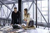 Two people are focused on a laptop, surrounded by papers and fabric samples on a large table. The setting is a modern office with large windows and a view of a building exterior.