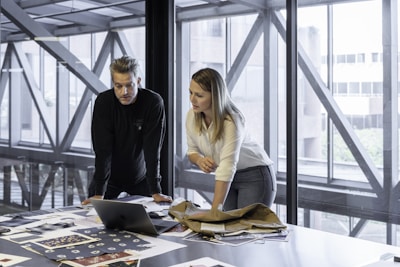 Two people are focused on a laptop, surrounded by papers and fabric samples on a large table. The setting is a modern office with large windows and a view of a building exterior.