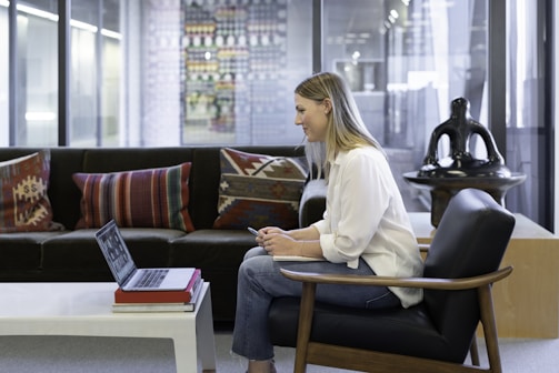 A person sits on a chair in a modern office environment, using a laptop placed on a white coffee table. The setting features a comfortable dark sofa with patterned cushions and a large decorative sculpture in the background. Glass partitions line the room, suggesting an open-layout office.