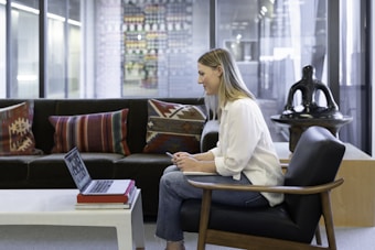 A person sits on a chair in a modern office environment, using a laptop placed on a white coffee table. The setting features a comfortable dark sofa with patterned cushions and a large decorative sculpture in the background. Glass partitions line the room, suggesting an open-layout office.