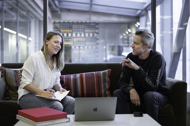 Two people are sitting on a sofa in a modern office setting, engaged in conversation. One is holding a notebook and pen, taking notes, while the other gestures with their hand. A laptop rests on a table in front of them along with a smartphone and some books. The room is well-lit with large windows and a background featuring a contemporary design.