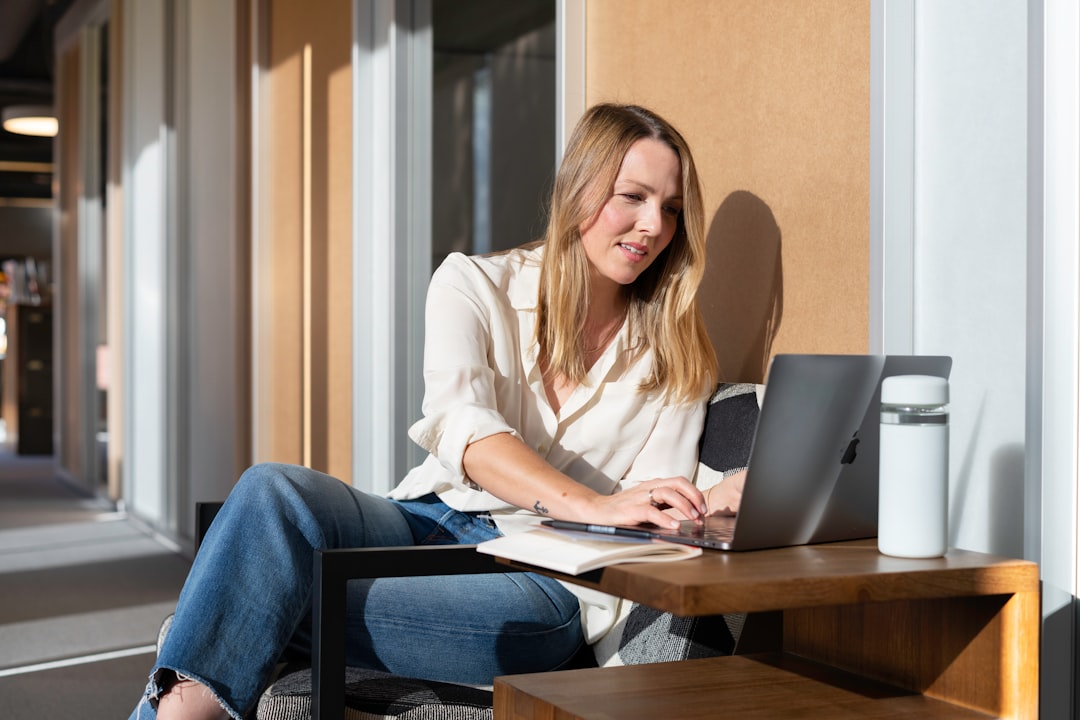 woman in white long sleeve shirt and blue denim jeans sitting on brown wooden table using, A salesperson working in an office at a laptop in the sun