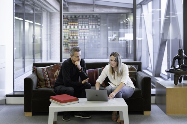 Two people seated on a sofa are engaged with a laptop on a coffee table. The setting appears to be an office or modern workspace with large windows and a background featuring an art display. A red folder is also on the table, and a decorative sculpture is nearby.