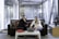 Two people seated on a sofa are engaged with a laptop on a coffee table. The setting appears to be an office or modern workspace with large windows and a background featuring an art display. A red folder is also on the table, and a decorative sculpture is nearby.