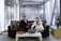 Two people seated on a sofa are engaged with a laptop on a coffee table. The setting appears to be an office or modern workspace with large windows and a background featuring an art display. A red folder is also on the table, and a decorative sculpture is nearby.