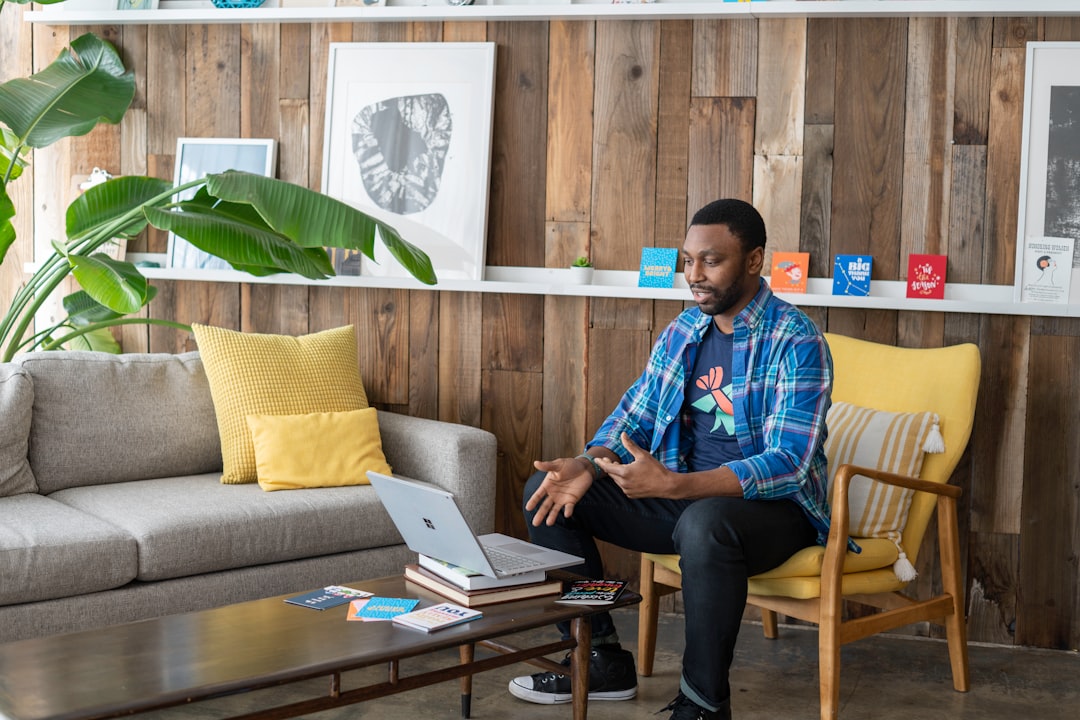 man in blue and white plaid dress shirt and black pants sitting on couch using macbook