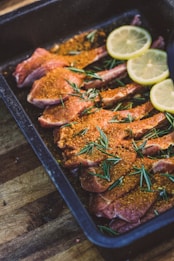 A black roasting pan contains several raw lamb chops seasoned with a reddish-orange spice mix. Sprigs of fresh rosemary are scattered on top of the meat, adding a touch of green. Slices of fresh lemon are placed at one end of the pan, contributing a hint of yellow contrast. The pan rests on a wooden surface, likely a kitchen countertop.