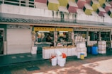 A market stall with a vibrant yellow sign displays various goods, including dry beancurd, noodles, and other provisions. The area is surrounded by white and blue plastic barrels, boxes, and containers. Above the stall, there is a building facade with a geometric pattern featuring yellow, pink, and red tones.