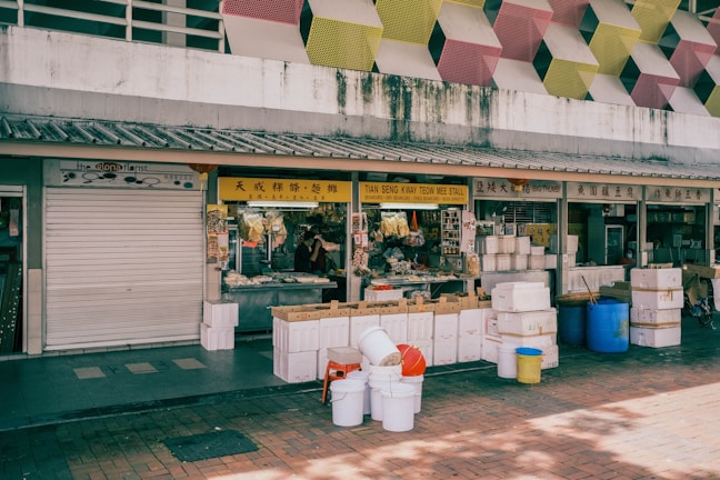 A market stall with a vibrant yellow sign displays various goods, including dry beancurd, noodles, and other provisions. The area is surrounded by white and blue plastic barrels, boxes, and containers. Above the stall, there is a building facade with a geometric pattern featuring yellow, pink, and red tones.