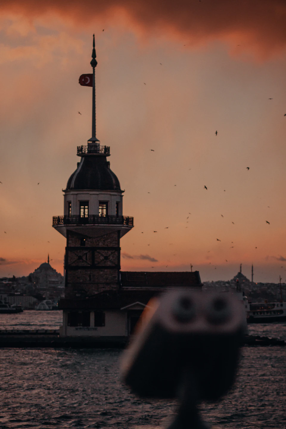 Tall white tower stands against orange sky near water's edge with flagpole on top
