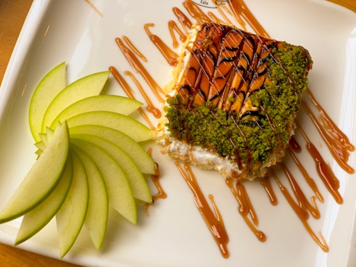 A piece of cheesecake is topped with a vibrant green pistachio crumble and drizzled with caramel and chocolate syrup. Next to it are thinly sliced green apple pieces arranged in a fan shape on a white rectangular plate.