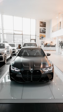 A sleek black car with a shiny, modern design is prominently displayed in a spacious and well-lit showroom. The showroom features large windows allowing natural light to pour in, highlighting the vehicle's features. Other cars are lined up in the background along the sides.