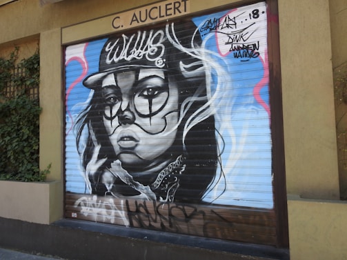 A mural painted on a metal shutter features a stylized portrait of a woman wearing a cap. The artwork incorporates bold, abstract shapes and lines around the face, with graffiti-style text above the figure.