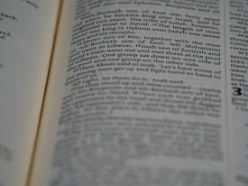 A page from a book is open, displaying text that appears to be from a biblical scripture. The text is in two columns and some sections are blurred due to the depth of field effect. The image focuses on a specific passage, with other parts of the page slightly out of focus.