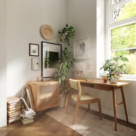 A cozy, well-lit corner of a room features a wooden desk and chair placed next to a large window with sunlight streaming in. The desk is adorned with a small potted plant and a vase of flowers, adding a touch of greenery. Nearby, a mid-century modern cabinet holds art supplies or books, with several frames and prints decorating the wall above. A variety of houseplants enhance the atmosphere, creating a serene and natural environment.