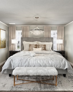 A luxurious bedroom with a large bed that has an upholstered headboard and is layered with multiple pillows and a soft throw. On either side of the bed, there are dark bedside tables with modern lamps. A plush, fur-covered bench sits at the foot of the bed on a textured area rug. The ceiling features an elegant crystal chandelier. Neutral-colored curtains frame two windows in the background, and a piece of abstract art adorns the wall.
