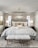 A luxurious bedroom with a large bed that has an upholstered headboard and is layered with multiple pillows and a soft throw. On either side of the bed, there are dark bedside tables with modern lamps. A plush, fur-covered bench sits at the foot of the bed on a textured area rug. The ceiling features an elegant crystal chandelier. Neutral-colored curtains frame two windows in the background, and a piece of abstract art adorns the wall.
