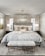 A luxurious bedroom with a large bed that has an upholstered headboard and is layered with multiple pillows and a soft throw. On either side of the bed, there are dark bedside tables with modern lamps. A plush, fur-covered bench sits at the foot of the bed on a textured area rug. The ceiling features an elegant crystal chandelier. Neutral-colored curtains frame two windows in the background, and a piece of abstract art adorns the wall.