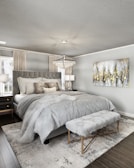 A modern bedroom features a large, upholstered bed with plush gray bedding. A fluffy throw blanket and multiple decorative pillows in varying textures complement the luxurious aesthetic. A soft, furry bench sits at the foot of the bed on a textured area rug. Twin nightstands with lamps flank the bed, and a large crystal chandelier hangs from the ceiling, adding elegance. A framed abstract painting with gold, gray, and white colors adorns the wall, and natural light filters through sheer curtains.