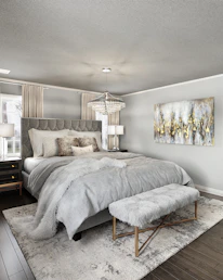 A modern bedroom features a large, upholstered bed with plush gray bedding. A fluffy throw blanket and multiple decorative pillows in varying textures complement the luxurious aesthetic. A soft, furry bench sits at the foot of the bed on a textured area rug. Twin nightstands with lamps flank the bed, and a large crystal chandelier hangs from the ceiling, adding elegance. A framed abstract painting with gold, gray, and white colors adorns the wall, and natural light filters through sheer curtains.