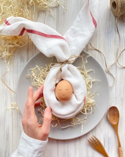 A creatively styled setting with an egg placed in the center of a white napkin folded to resemble rabbit ears. The napkin is tied with twine and the egg is nestled on a bed of straw on a gray plate. The egg has a cute, drawn-on face with closed eyes and a smile, suggesting the appearance of a sleeping baby. A hand gently touches the napkin, adding a sense of care. The scene is surrounded by various kitchen utensils and additional straw spread on a light wooden table.