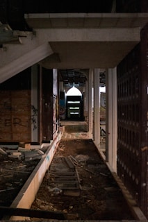An abandoned, partially constructed building with exposed concrete beams and columns. The floor is littered with debris, broken planks, and remnants of a makeshift railway track. Graffiti is visible on the walls, and a faint light filters through openings, creating a dimly lit and eerie atmosphere.