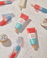 A playful arrangement of colorful popsicles, cupcakes, and two tubes of a product labeled 'not pot' displayed on a surface sprinkled with glitter and star-shaped confetti, creating a whimsical and festive atmosphere.