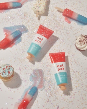 A playful arrangement of colorful popsicles, cupcakes, and two tubes of a product labeled 'not pot' displayed on a surface sprinkled with glitter and star-shaped confetti, creating a whimsical and festive atmosphere.