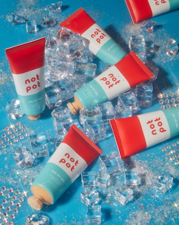 Several tubes labeled 'not pot' are scattered on a blue surface alongside acrylic ice cubes and glitter, creating a colorful and playful composition. The tubes have red and teal colors with white text.