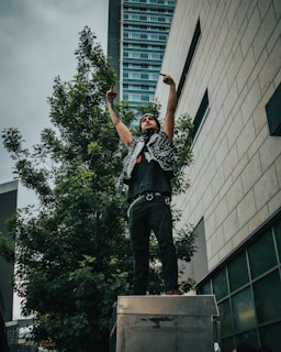 A person stands on a metal surface, raising both middle fingers. They wear a sleeveless, studded leather vest, black pants, and a bandana on their head. The background includes a tall building with a modern design and a large tree.