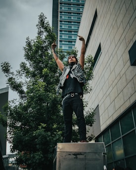 A person stands on a metal surface, raising both middle fingers. They wear a sleeveless, studded leather vest, black pants, and a bandana on their head. The background includes a tall building with a modern design and a large tree.